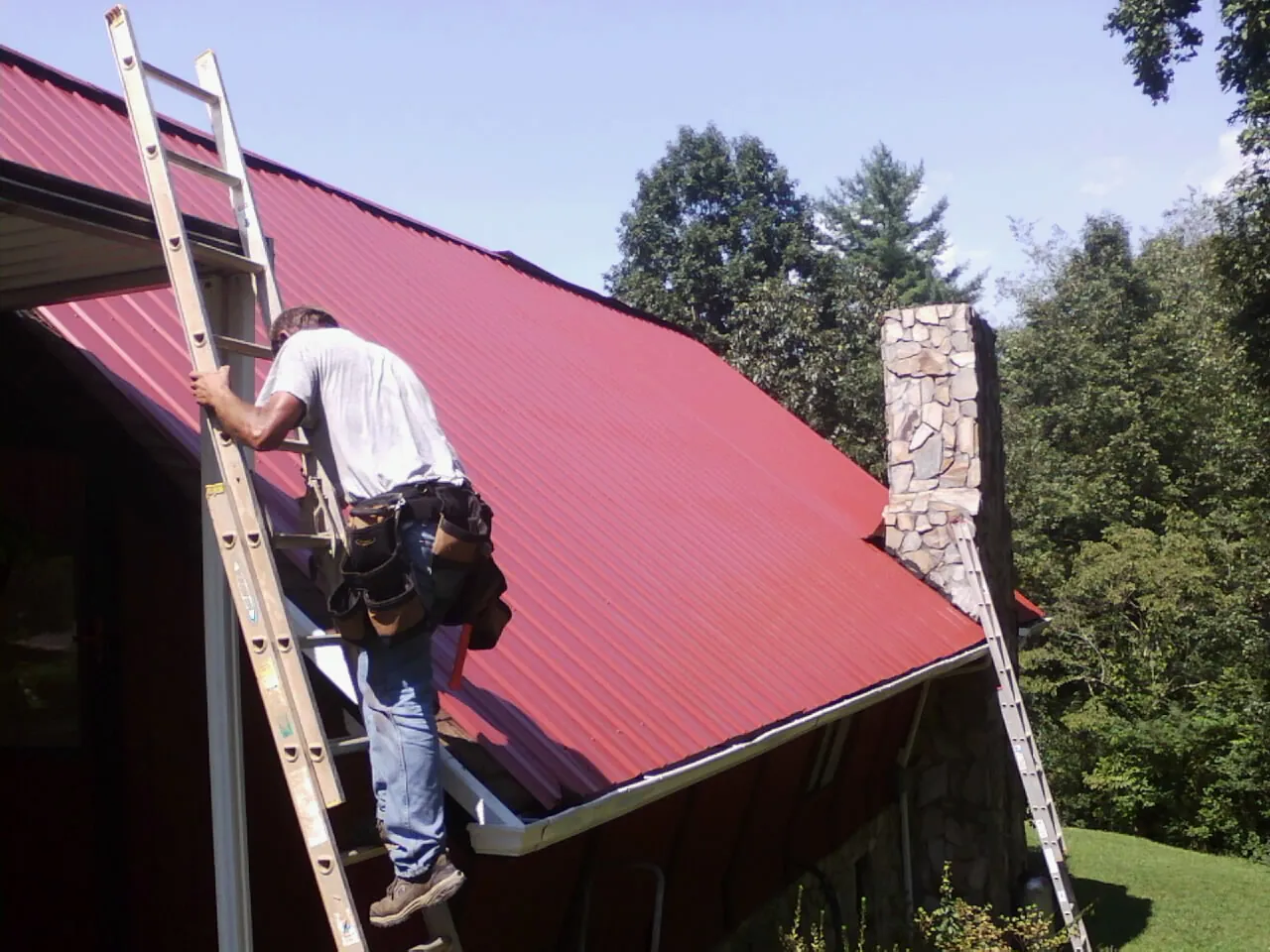 Roofer on a ladder beside a red metal roof and stone chimney — Soffit & Fascia Repair by Roofing Brothers in Addison